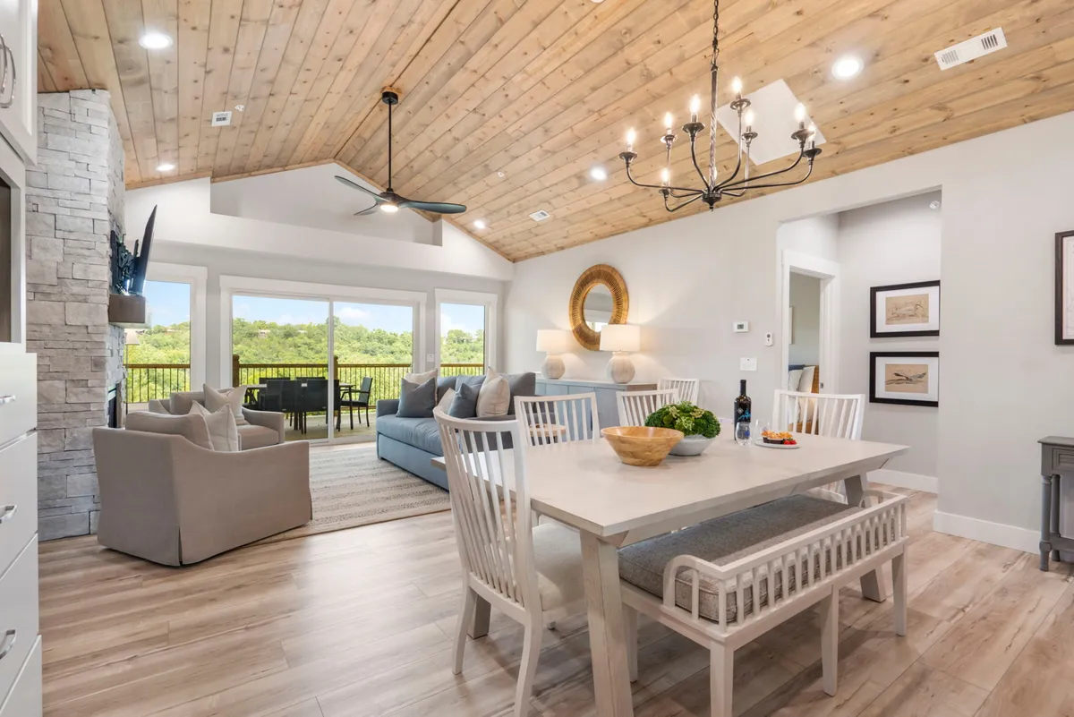 The image showcases a warm, inviting open-concept living and dining space filled with natural light from large windows. A stylish dining table sits beneath a modern chandelier, set for family gatherings or celebratory meals. A plush sofa offers a cozy spot for relaxing or reading, all framed by scenic outdoor views visible through the windows.