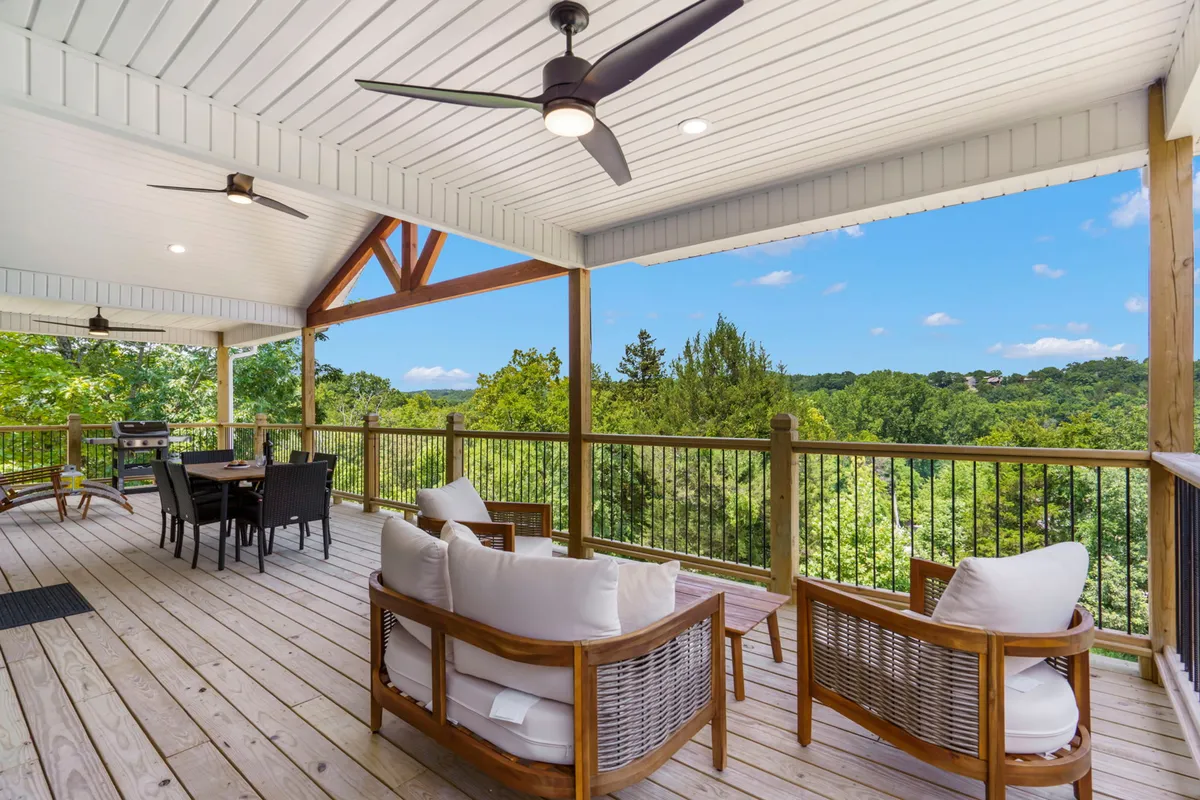 A spacious deck with a dining table and comfortable seating overlooks scenic hills and trees. Ceiling fans offer comfort during warm days, and the space looks ready for both morning coffees and evening gatherings. The open view and ample seating invite guests to relax and enjoy the outdoors year-round.