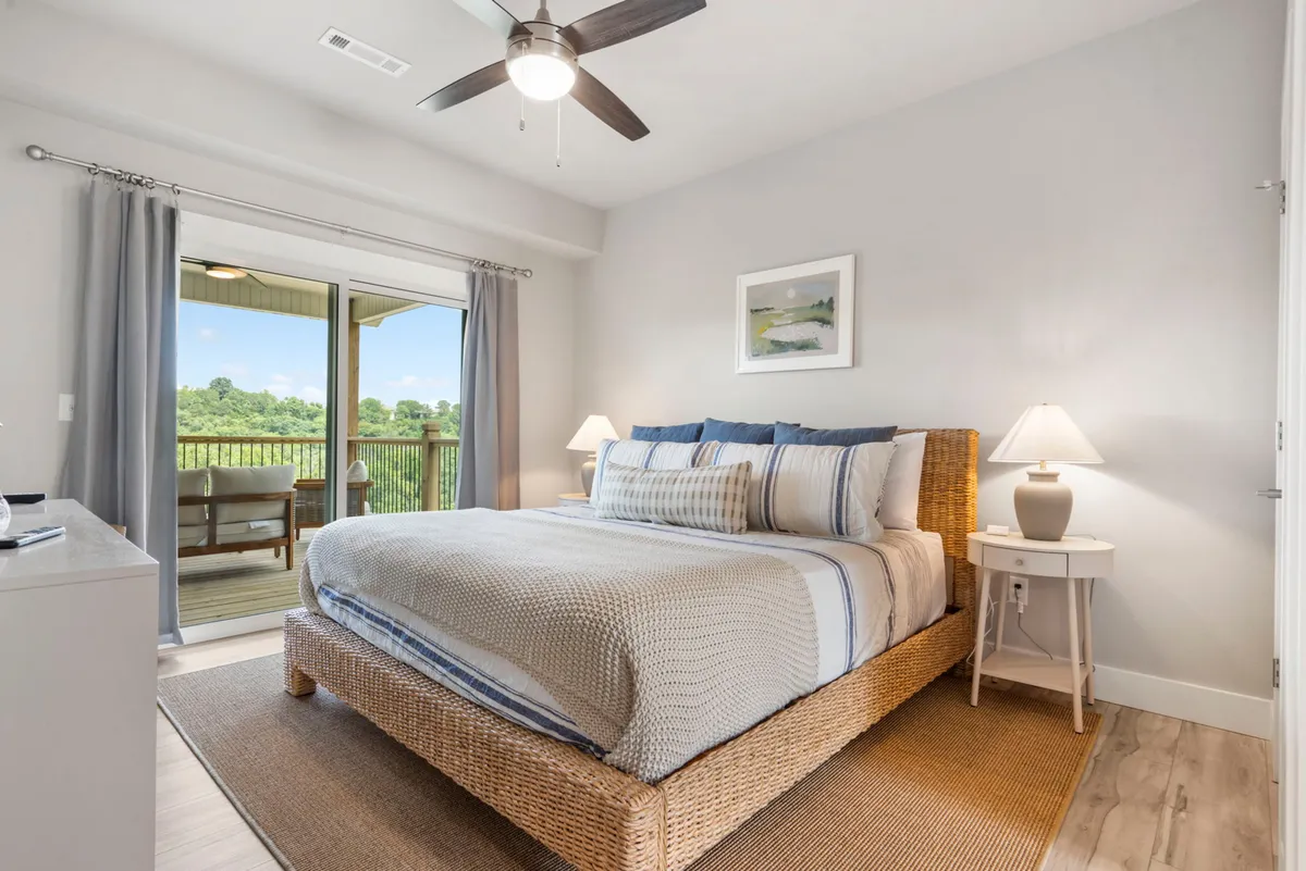 A tranquil king bedroom featuring a bed with decorative pillows, a sliding glass door leading to a balcony, a nightstand with a lamp, and artwork on the wall. Natural light streams into the room, creating a relaxing and inviting ambiance.