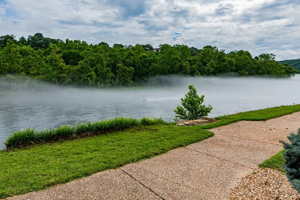 A serene river winds through lush trees, with soft morning fog drifting above the water and a rustic pathway running alongside the shoreline. Gentle sunrise reflections shimmer on the water’s surface, and the natural setting suggests opportunities for boating and peaceful strolls.