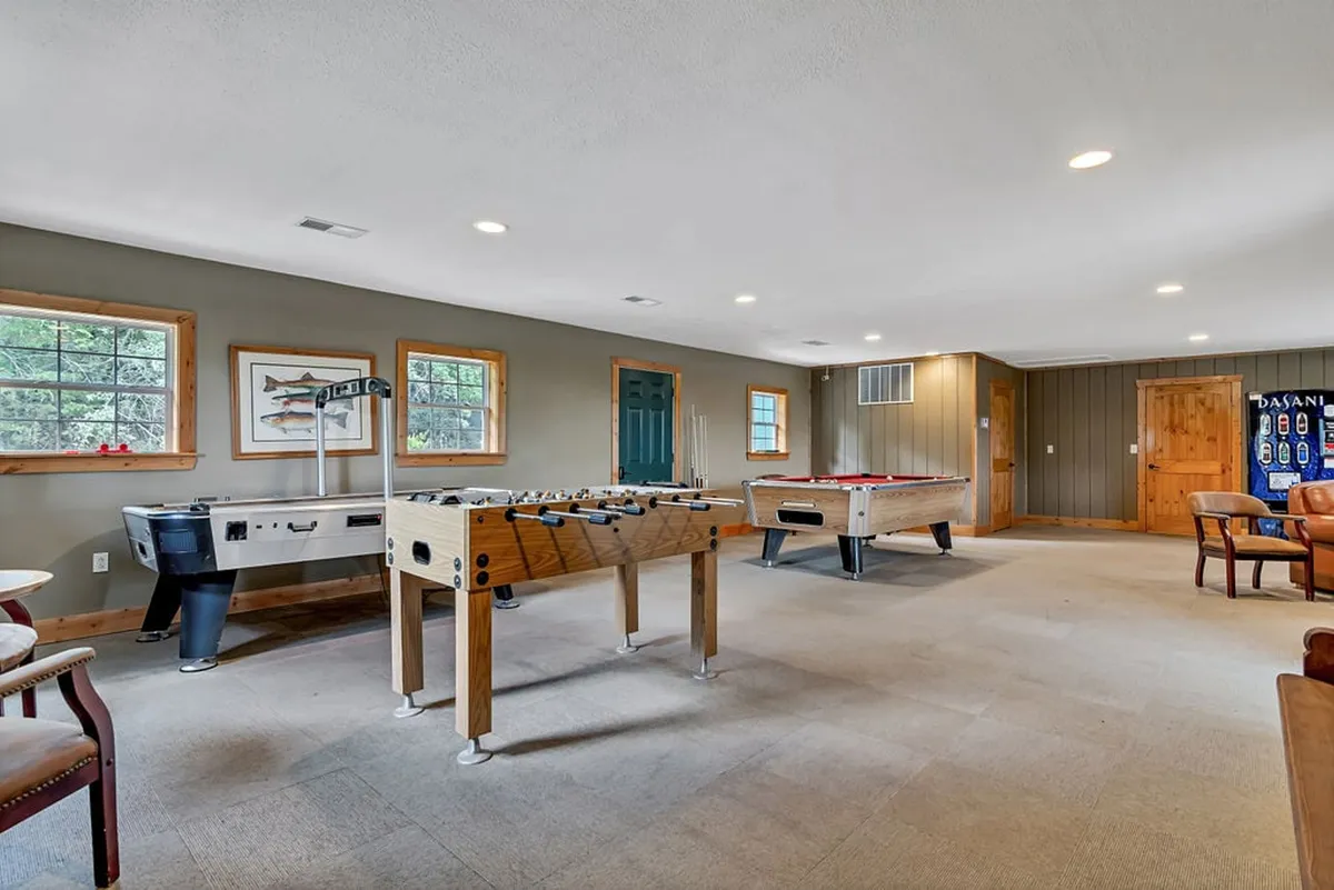 A spacious playroom featuring a foosball table, pool table, and seating area by large windows, all bathed in natural light. The setup is energetic and inviting, encouraging group activities and family fun.