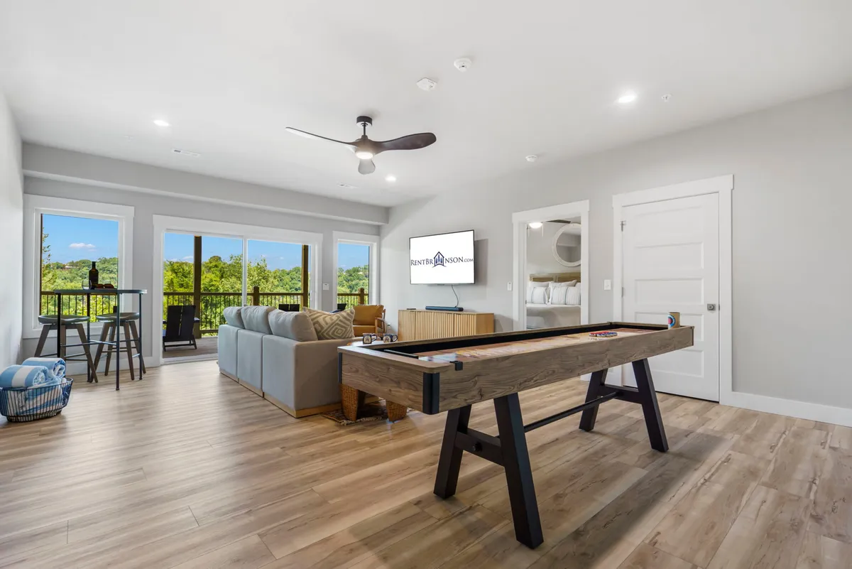 A lively lower-level living space featuring a shuffleboard table, comfortable sofa, large windows letting in natural light, and a TV mounted on the wall. The arrangement emphasizes social interaction and fun family activities.